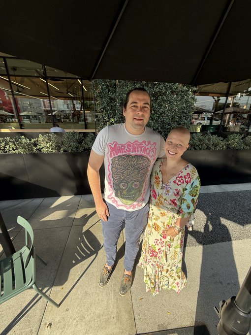 A man and woman posing together outdoors on a sunny day; the man wears a white graphic t-shirt and blue pants, while the woman wears a floral dress.