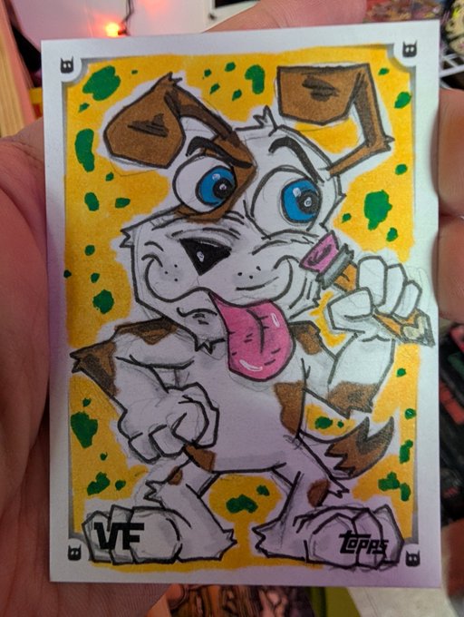 A hand holding a hand-drawn sketch card featuring a cartoon dog with brown patches, blue eyes, and its tongue hanging out, signed 'VF' and 'TOPPS' on a yellow background with green spots.