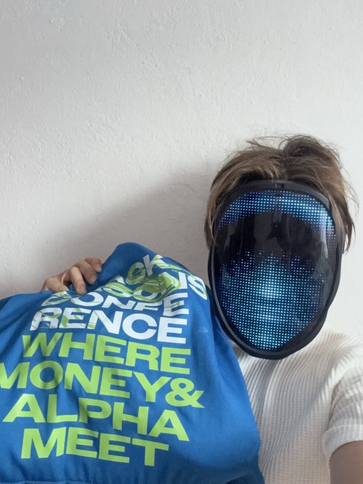 A person wearing a black LED mask with blue lights forming a face pattern, holding up a blue shirt that reads 'CONFERENCE WHERE MONEY & ALPHA MEET'.