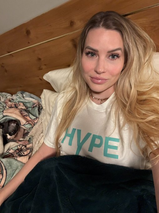 A young woman with long blonde hair wearing a white t-shirt with 'HYPE' printed in teal letters poses for a selfie on a bed with a wooden headboard, with a small dog visible under a blanket.