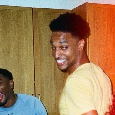 A photograph of a smiling young man wearing a yellow shirt, with another partially visible person beside him in front of wooden cabinets.