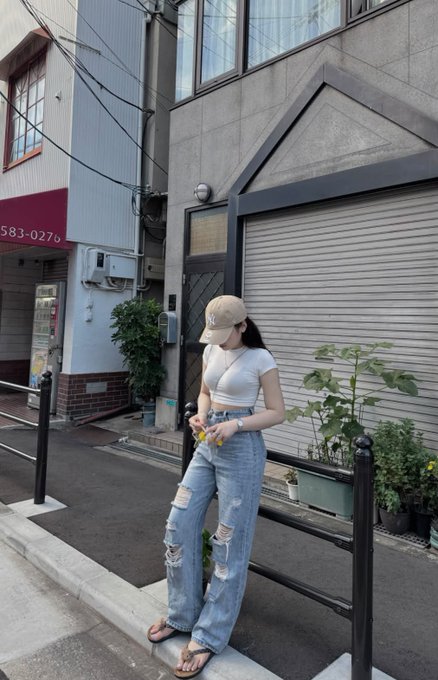A young woman wearing a beige NY Yankees cap, white crop top, and distressed light-wash jeans standing on a sidewalk in front of a building with a metal roll-up door, holding a small snack.