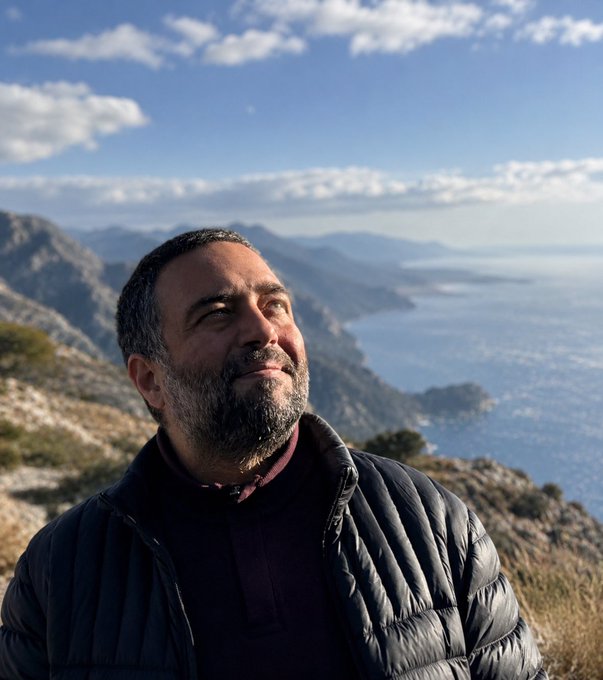 The image features a man standing at the top of a mountain with his back turned towards the camera. He appears to be looking out over the ocean, taking in the beautiful view. The surrounding landscape includes mountains and water, creating an impressive backdrop for this picturesque scene.