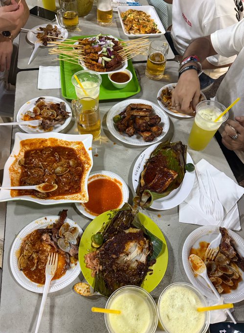 A table full of various dishes including satay skewers, clams in sauce, fish, rice, and drinks, with people seated around it.