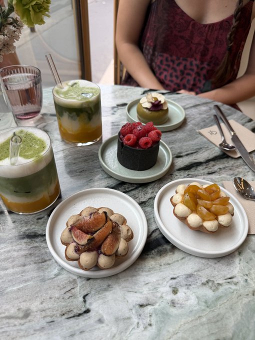 A marble table holds an assortment of pastries including tarts topped with figs, raspberries, and pears, alongside layered green matcha drinks with glass straws, with a person in a leopard-print top seated across the table.