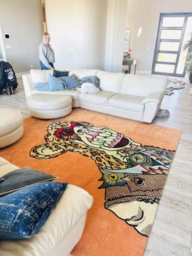 A living room features a large orange area rug with a colorful leopard design, accompanied by a white sectional sofa and cream ottomans.