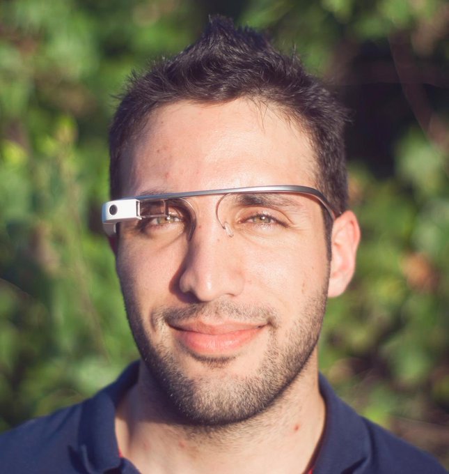 The image features a man wearing glasses with a smile on his face as he looks directly at the camera. The man appears to be a young adult, possibly a teenager or a young man, who seems happy and content while wearing the glasses.