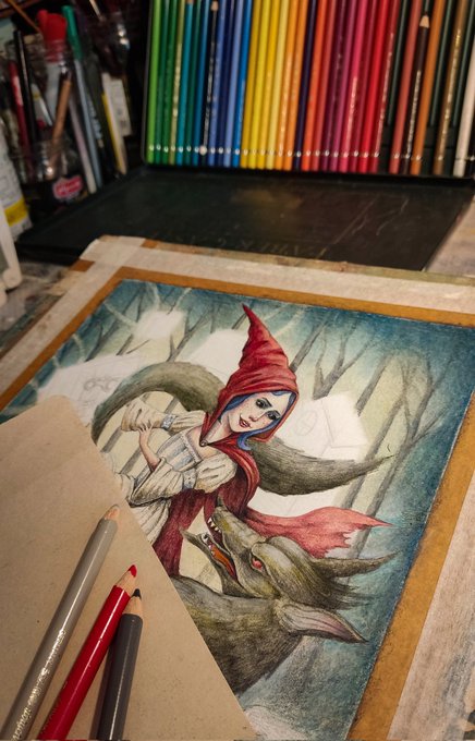 A colored pencil drawing of a hooded character with a wolf, surrounded by colored pencils, in progress on a desk.