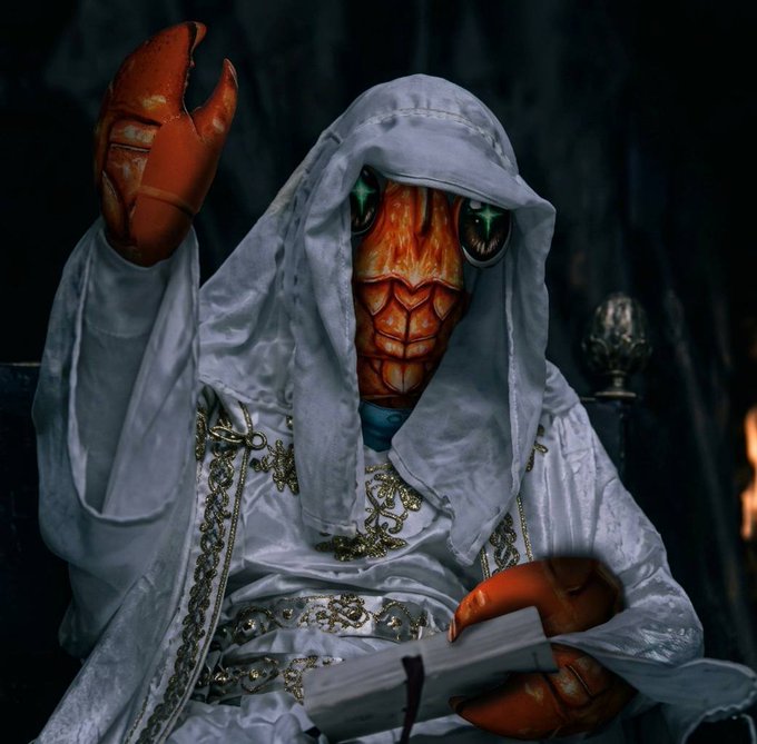 A person in elaborate cosplay featuring a white hooded robe with gold detailing and an orange crustacean-like face mask with green eyes, holding a book and raising one claw.