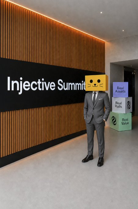 A person in a gray suit with a yellow cat emoji face stands in a modern lobby next to an 'Injective Summit' sign and stacked blocks labeled 'Real Assets', 'Real Rails', and 'Real Value'.