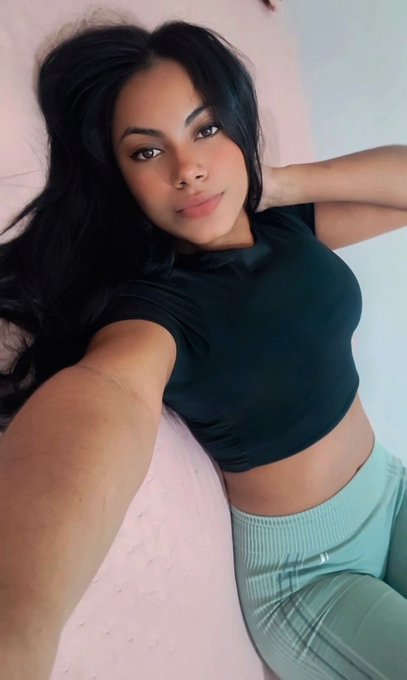 A selfie-style photo of a young woman with long black hair, wearing a black cropped t-shirt and light green pants, posing against a textured pink and white background.