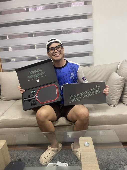 A smiling young man wearing glasses, a backwards cap, and a blue sports jersey sits on a couch holding up a 'Joysent' branded box that contains a red and black gaming paddle.