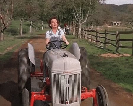 A woman in a blue dress drives a gray tractor with red wheels down a dirt road, with another person visible in the background.