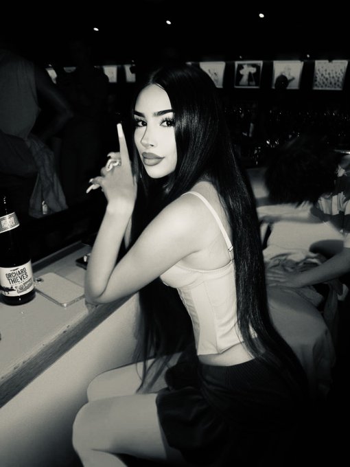 A black and white photo of a young woman with long dark hair sitting at a bar with a bottle of Michelob Ultra, pointing her finger toward the camera while looking over her shoulder.