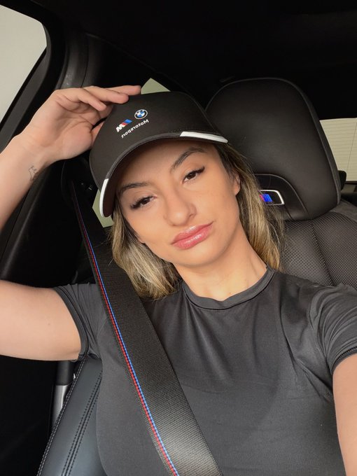 A woman wearing a black BMW M Motorsport cap and a gray t-shirt sits in the driver's seat of a car with the seatbelt fastened, posing for a selfie.