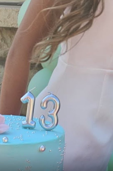 A young girl in a white dress stands beside a light blue birthday cake adorned with silver "13" candles and pearl-like decorations, celebrating her 13th birthday, which the post joyfully connects to the XRP community's hope for the price target of $13.