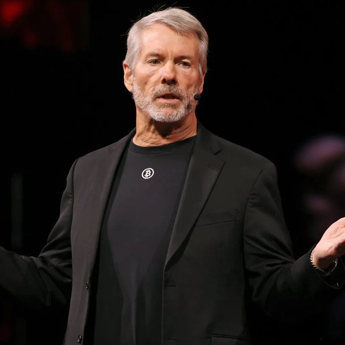 An older man with grey hair and beard wearing a black blazer over a Bitcoin logo t-shirt, appearing to give a presentation.