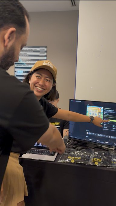 A smiling woman wearing a tan "Hokuriku" cap points excitedly at a computer screen showing what appears to be an NFT marketplace, likely Rarible, while a man in a black shirt leans in beside her at what looks like a conference booth with branded merchandise on the table. The moment captures the excitement of successfully pulling off an NFT trade together, matching the post's celebration of their joint accomplishment.