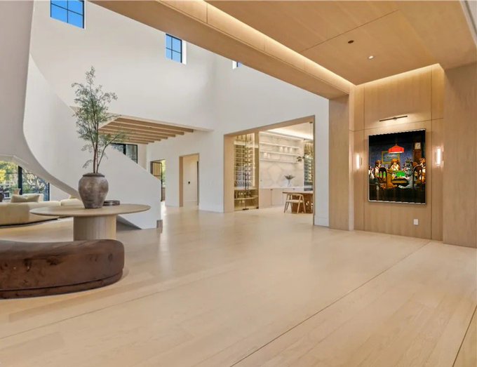 A spacious, modern living room with light wood floors, white walls, a curved staircase, and minimalist furniture, featuring an olive tree in a large vase and a colorful painting on the right wall.