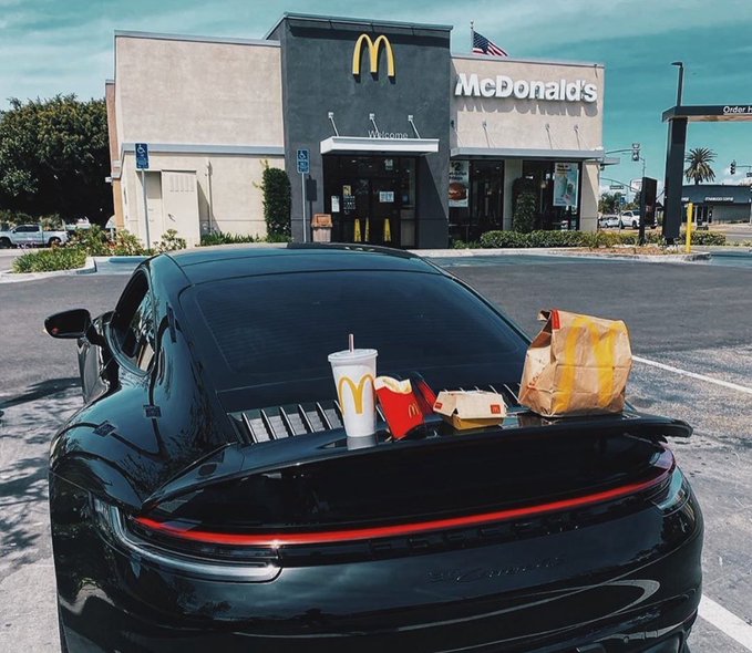 The image shows a black car with a meal from Mcdonald's placed on its back bumper, which includes two Mcdonald's sandwiches as well as two cups of coffee. The text in the accompanying post seems unrelated to the actual meal but might be providing additional context or information about something else. This could be related to the post that says "Year 2035: I tell my kids the story of when I bought $BTC at $60k Saturday afternoon, Porsche, McDonald's, Family." It may be a humorous story or an interesting piece of information unrelated to the meal on the car.
