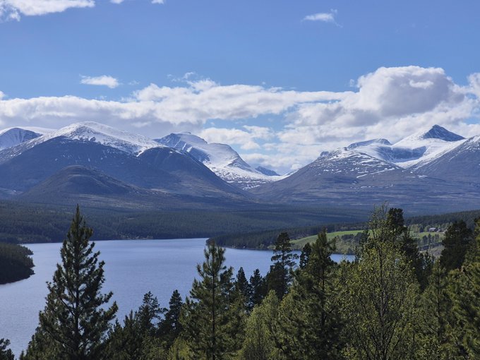 A serene mountain landscape featuring snow-capped peaks, a tranquil lake, and evergreen trees under a partly cloudy sky, complementing the post's theme of a new nature photography drop titled "From a Distance" being released as a 1/1 original.