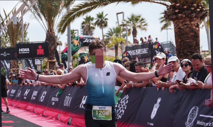A runner wearing bib number 1208 (Artem) crossing a finish line with arms outstretched in triumph, surrounded by cheering spectators, with HOKA and "Fly Human Fly" branding visible.