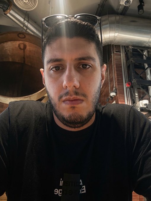 A close-up selfie of a bearded man wearing a black t-shirt with sunglasses resting on his head, photographed in an industrial-looking indoor setting.
