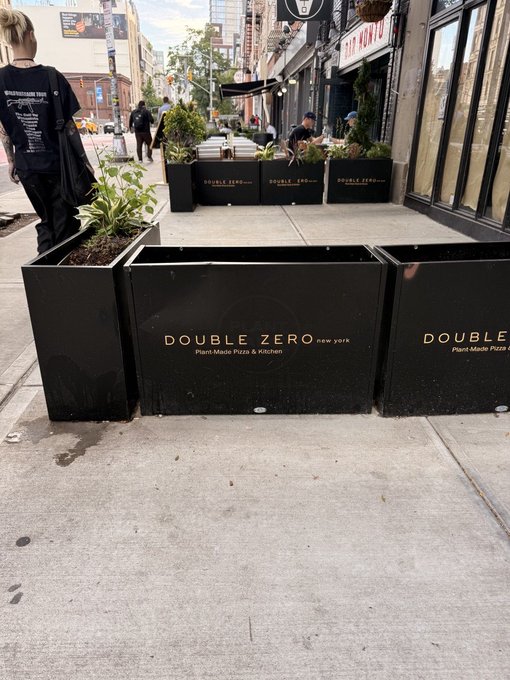 A street-level view of an outdoor dining setup with black metal planters labeled 'DOUBLE ZERO new york Plant-Made Pasta & Kitchen' blocking a sidewalk, with pedestrians and storefronts including 'NEW YORK' signage in the background.