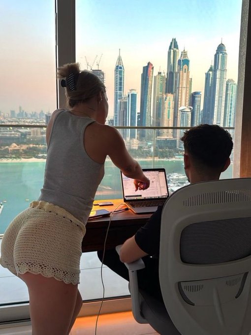 Two people look at a laptop together by a window overlooking a stunning city skyline with skyscrapers and water, while one leans in pointing at the screen, matching the "good afternoon legends who's active?" vibe with their afternoon work session in a high-rise setting.