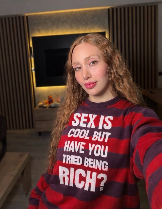 A young woman with long curly blonde hair takes a selfie wearing a red and dark blue striped sweater that reads 'SEX IS COOL BUT HAVE YOU TRIED BEING RICH?'.