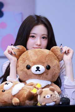 A young woman with long dark hair posing with a brown Rilakkuma teddy bear, holding it in front of her.