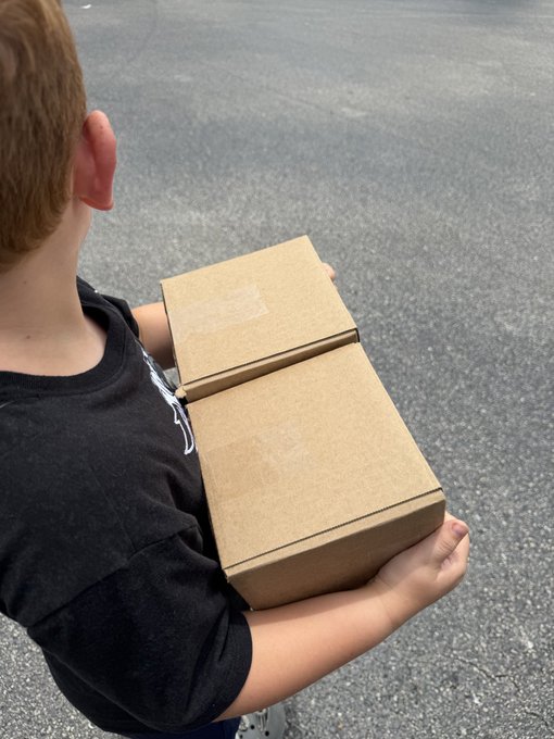 A young person in a black t-shirt holding two stacked cardboard boxes outdoors on asphalt pavement.