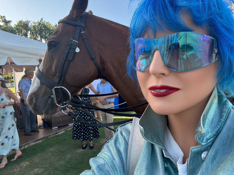 A selfie showing a person with bright blue hair wearing large iridescent shield sunglasses and red lipstick, posing next to a brown horse wearing a bridle at an outdoor event.
