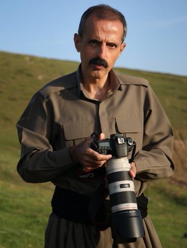 A portrait of a middle-aged man with a mustache wearing an olive-green shirt, standing in a grassy field while holding a DSLR camera with a large telephoto lens.