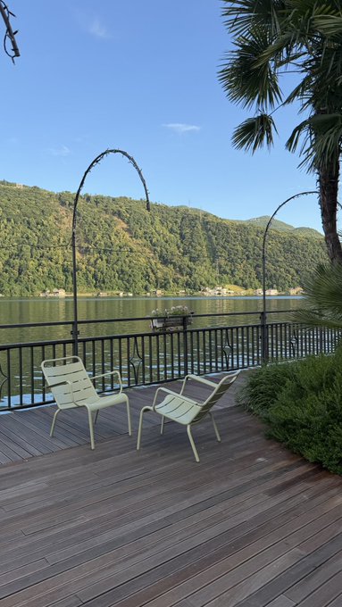 The image shows a wooden deck with two chairs, one white chair and one green chair, facing each other. There are also some trees visible near the deck. The photo has been edited to include a caption that reads "Life when you comfy hold $LFI".