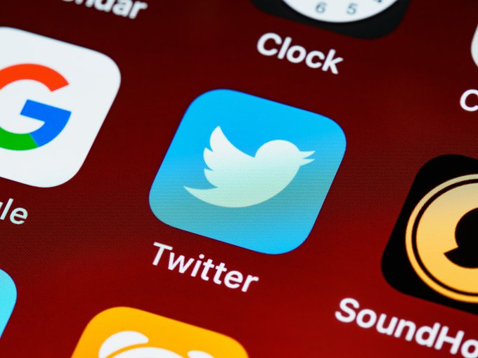 A close-up photo of a smartphone screen displaying the Twitter app icon alongside other apps like Google, Calendar, Clock, and SoundHound.