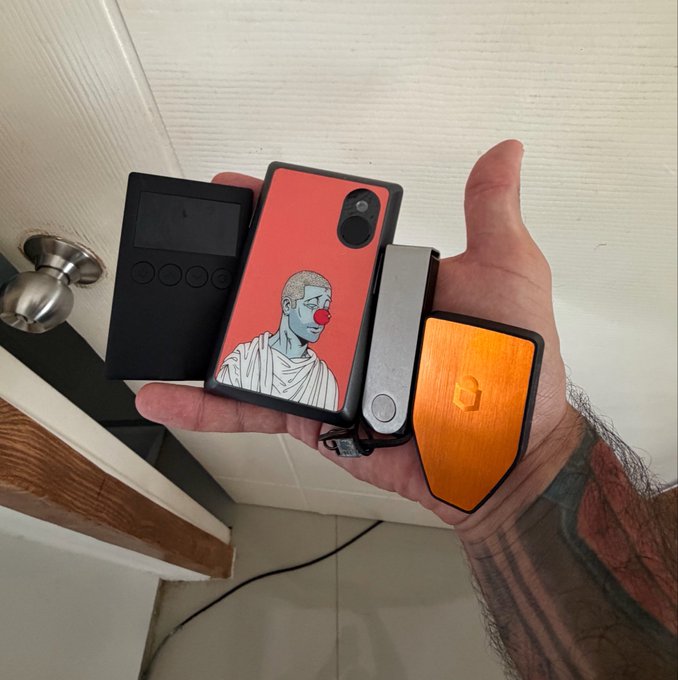 A hand proudly displays an arsenal of crypto hardware wallets, featuring a black device with buttons, a red phone-style wallet adorned with a classical Roman bust and red clown nose sticker, a silver rectangular wallet, and an orange metallic wallet, all set for upcoming reviews.
