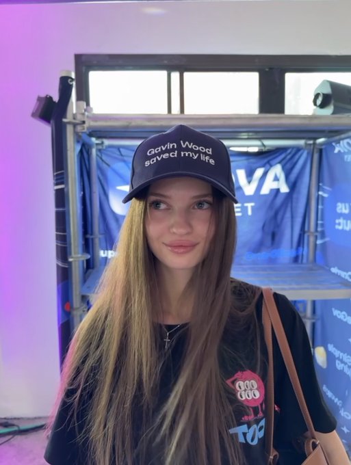 A young woman with long brown hair poses at what appears to be a crypto convention, wearing a navy cap that reads "Gavin Wood saved my life" — a nod to the Ethereum co-founder. Behind, a partially visible banner reads "AVA" and "...ET," consistent with the post's Ethereum-focused crypto lore, where she cites Ethereum as her first buy and biggest bag.