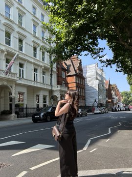 A woman in a black dress stands on a sunny city street lined with classic white buildings and trees.