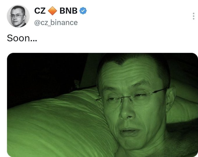 A screenshot of a tweet from CZ (@cz_binance) saying 'Soon...' above a green-tinted nighttime photo of him lying on a pillow.