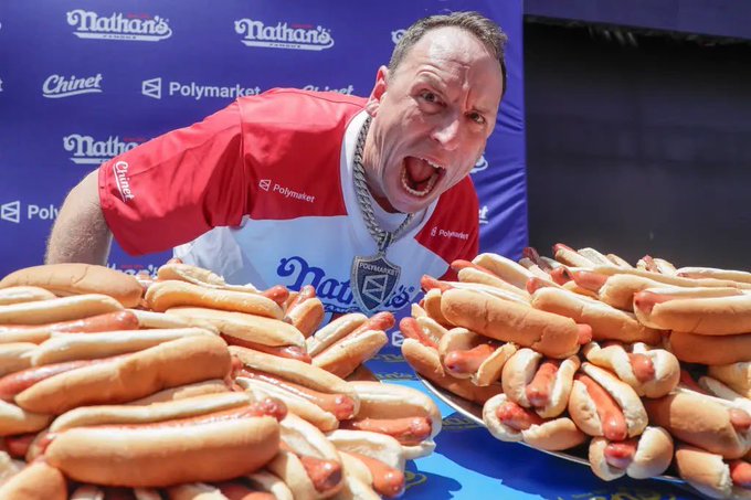 A victorious Joey Chestnut yells triumphantly behind massive platters piled high with hot dogs at Nathan's Famous Hot Dog Eating Contest, sponsored by Chinet and Polymarket, celebrating his record-setting win while sporting a large Polymarket championship chain.