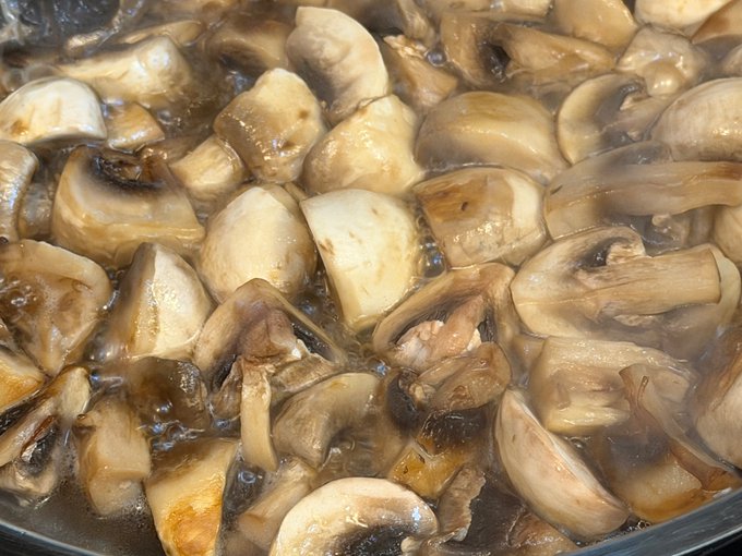 A close-up shot of sliced mushrooms simmering in a brown sauce or broth in a pan.