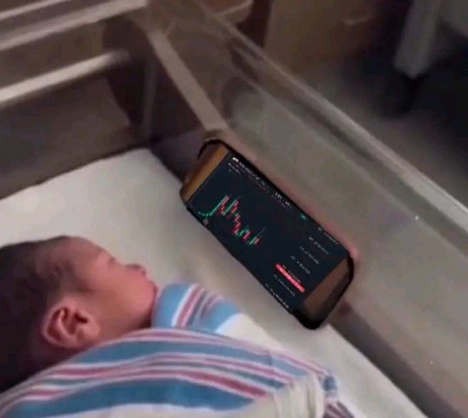 A baby lies in a crib wrapped in a striped blanket, with a smartphone propped up displaying a green candlestick stock or crypto trading chart.