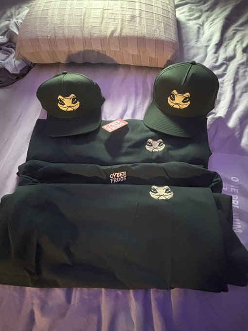 Black 'Cyber Frogs' streetwear collection displayed on a bed, including two caps with angry frog face logos and matching apparel with frog emblems.