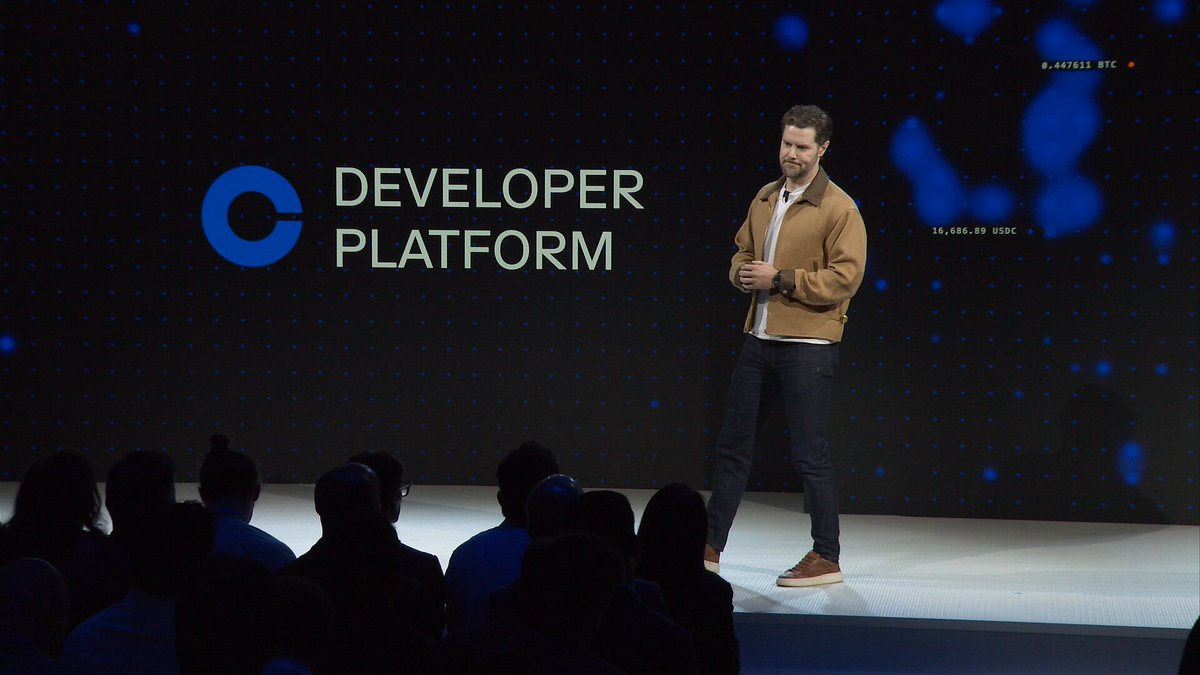 The image features a man standing on stage at a conference, giving a speech about the Coinbase Developer Platform. The platform is being introduced as a single access point for building with wallet infrastructure, payment capabilities, trading systems, and stablecoin issuance. The speaker is addressing an audience of people who are seated in front of him, indicating that he is speaking to them directly. The image also includes text that reads "Developer Platform" and "Welcome to the brand new Coinbase Developer Platform".