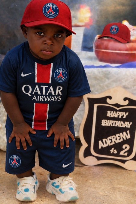 The image features a young boy wearing a soccer uniform with a red hat and blue shorts, standing next to a sign that says "Happy Birthday Adereen 1-2" and has an Arabic inscription. The background of the photo shows a dining table with various items on it, including a bowl and a cup.