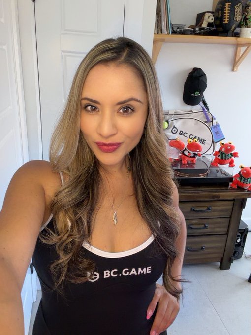 A woman with long blonde hair wearing a black 'BC.GAME' top poses for a selfie indoors, with BC.GAME merchandise and red bull figurines visible in the background.