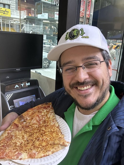 A smiling man wearing a white cap with a logo and a green shirt holds up a large slice of cheese pizza on a paper plate inside a pizzeria, with an ATM visible in the background.
