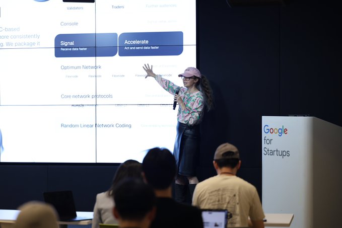 A presenter wearing a pink cap and floral shirt points toward a large screen displaying technical content about "Optimum Network" and "Core network protocols" with "Signal" and "Accelerate" buttons, while a podium to the right reads "Google for Startups." The setup reflects the post's focus on Muriel Medard explaining her networking technology at a startup event in Seoul, where she discusses optimizing the speed at which data moves between validators.