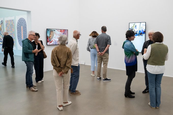A group of people in casual clothing viewing artwork on screens mounted in the white-walled gallery space of a museum or art exhibition.
